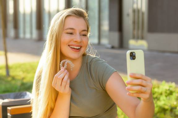 Frau macht Selfie und zeigt transparenten Zahnschienen-Aligner im Freien.